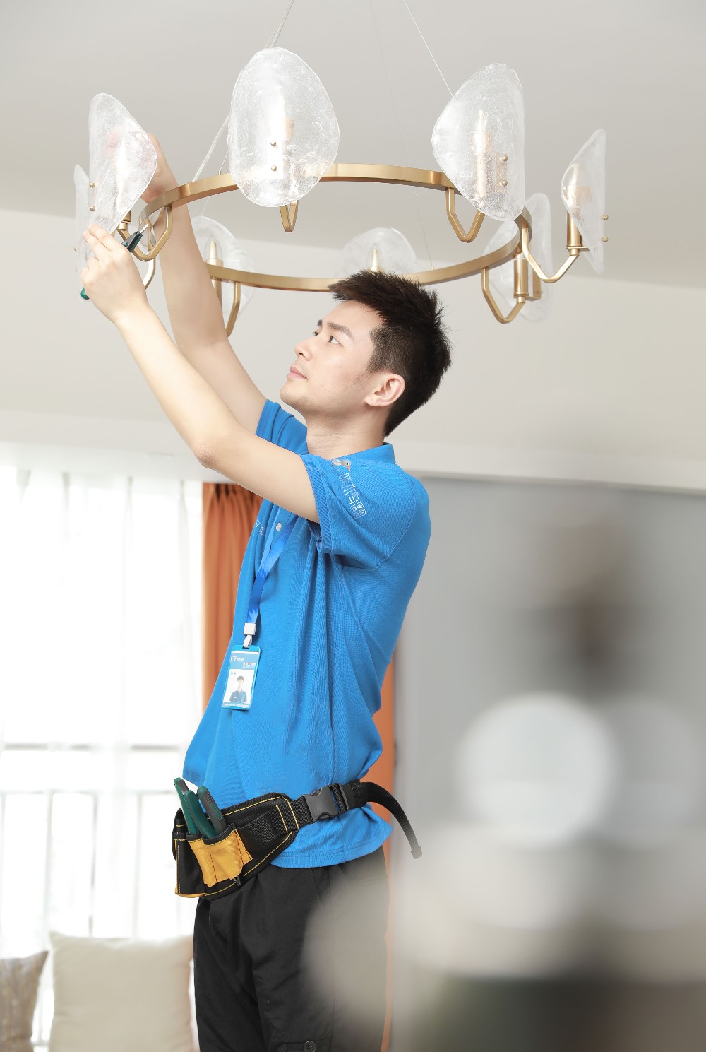 明亮的居家客厅环境中，身穿蓝色制服的专业维修师傅佩戴工具腰带，手持螺丝刀，正在专注安装一盏金色金属框架搭配不规则透明灯罩的圆形吊灯。画面展现专业上门灯具安装服务